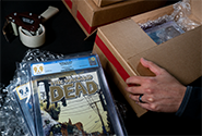 A CGC representative packages certified comic books in a cardboard box with void-filling materials.